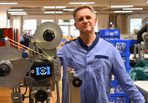 A man in a blue work uniform stands next to a machinery setup in a production environment.