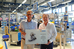 Two men are holding a certificate in a production hall, surrounded by machines and workstations.
