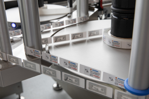 A labeling machine with multiple rollers that applies labels to a conveyor belt surface. The labels display various information.