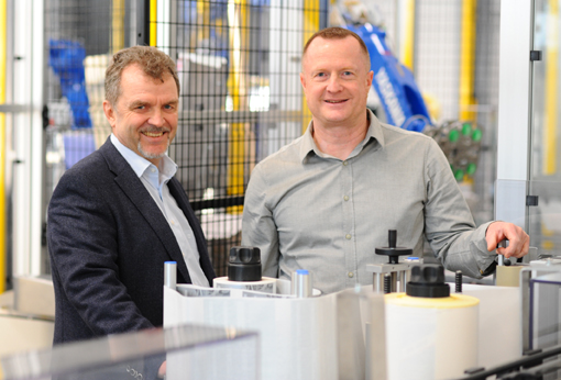Two men are standing in a modern production environment, surrounded by machines and equipment, smiling at the camera.