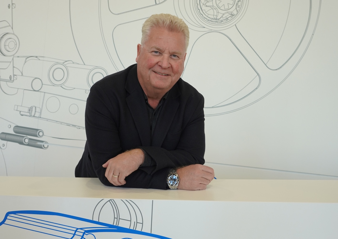 A man with blonde hair in a black blazer leans against a white table, smiling, in front of a sketched background graphic.