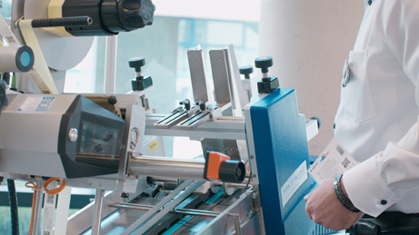 A person is operating a modern labeling machine equipped with various tools and a blue control panel.