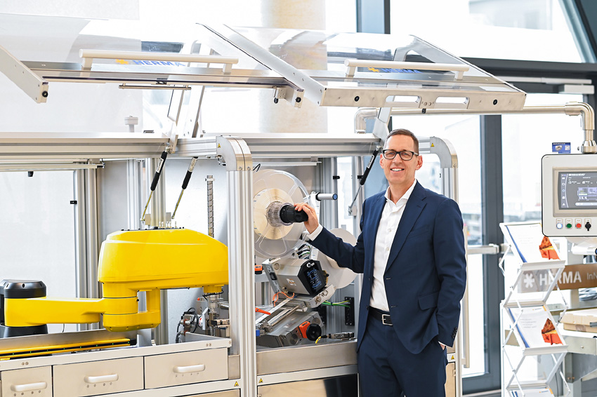 A man in a suit stands next to a modern machinery setup with yellow and silver components designed for industrial applications.