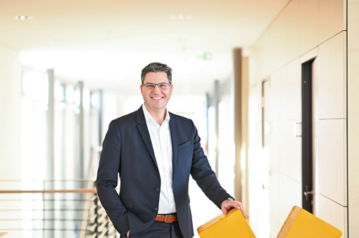 A man in a suit stands in a modern hallway, smiling as he holds onto the back of a yellow chair.