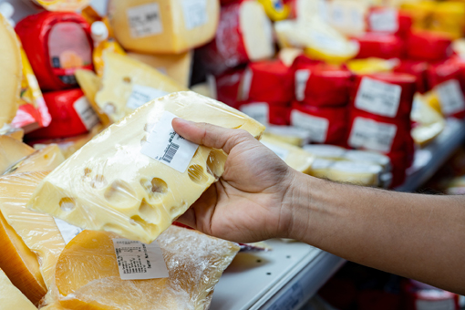 A hand is holding a piece of cheese with holes, while various types of cheese are resting on a shelf in the background.
