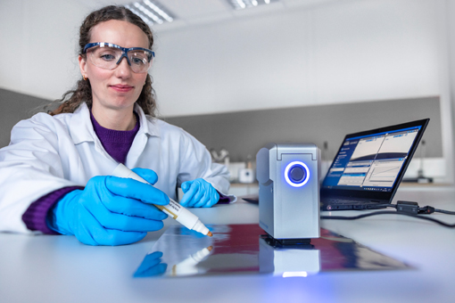 A person in a lab coat and safety goggles is using a marker to write on a shiny surface, while a laptop sits next to them.