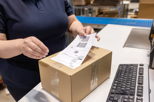 A person is sticking a shipping label onto a brown box while working at a desk with a keyboard.