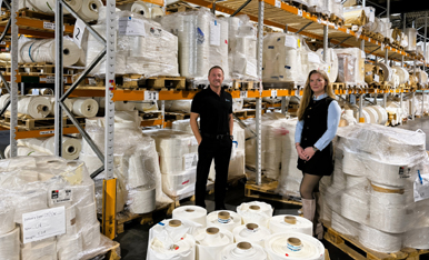 Two people are standing in a warehouse filled with large rolls of fabric or yarn, surrounded by shelves full of similar materials.