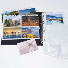 Ein Fotoalbum mit mehreren Seiten, die Landschafts- und Architekturaufnahmen zeigen, sowie leere Klarsichthüllen und eine Postkarte.