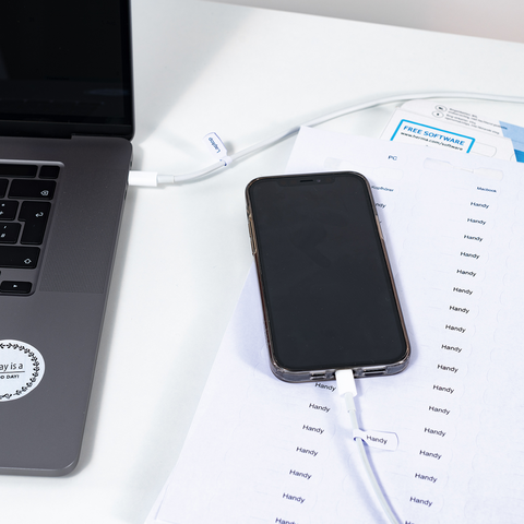 Ein Laptop und ein Smartphone liegen auf einem Tisch, verbunden durch ein Ladekabel. Neben dem Smartphone befinden sich ausgedruckte Dokumente.