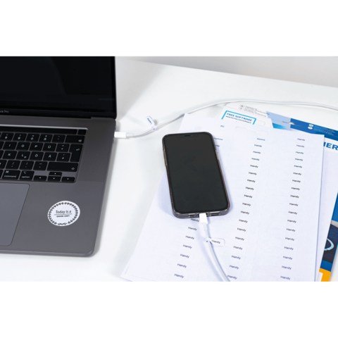 Ein Laptop und ein Smartphone liegen auf einem Tisch, verbunden durch ein Ladekabel. Neben dem Smartphone befinden sich ausgedruckte Dokumente.