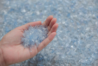 A hand holds a handful of blue, transparent plastic particles, surrounded by more similar particles on a surface.