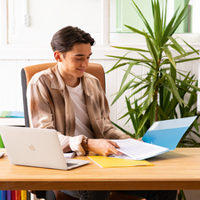 Ein junger Mann sitzt an einem Schreibtisch, arbeitet mit einem Laptop und blättert durch Unterlagen in einer hellen, modernen Umgebung.