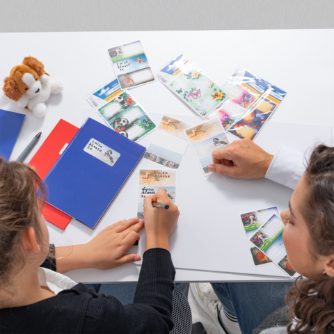 Hände halten ein gelbes Notizblatt mit einem Cartoon-Motiv, umgeben von Schulmaterialien, einem Buch und bunten Aufklebern.