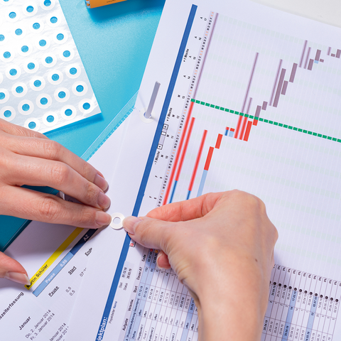 Hands place a round sticker on a document with charts and tables, while a pack of stickers is visible in the background.