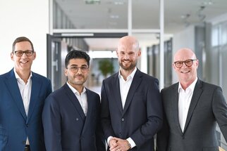 Four men in suits stand in a modern office building. They are smiling and posing cheerfully for the camera.