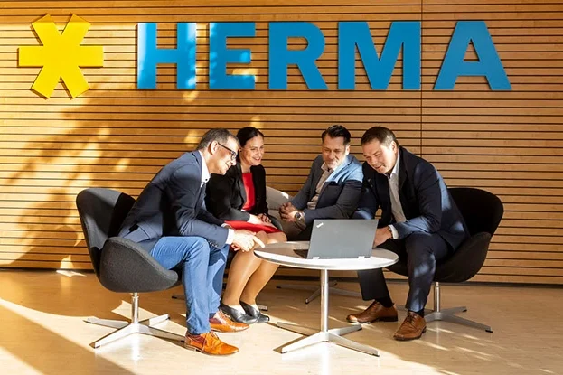 View of green treetops and a meeting of three people in a modern office with the sign "HERMA" in the background.