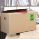 Kartonbox auf einem Tisch mit einem roten Heft und einem grünen "TOP"-Schild. Im Hintergrund sind Bücherregale sichtbar.