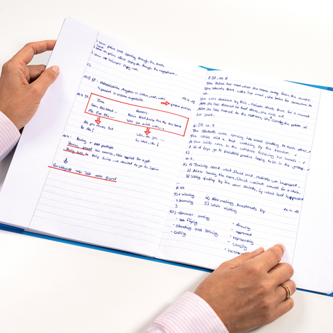 Hands hold an open notebook filled with handwritten notes and markings in blue and red ink.