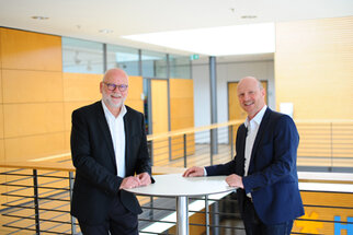 Two men are standing at a table in a modern office building. Both are wearing suits and smiling at the camera.
