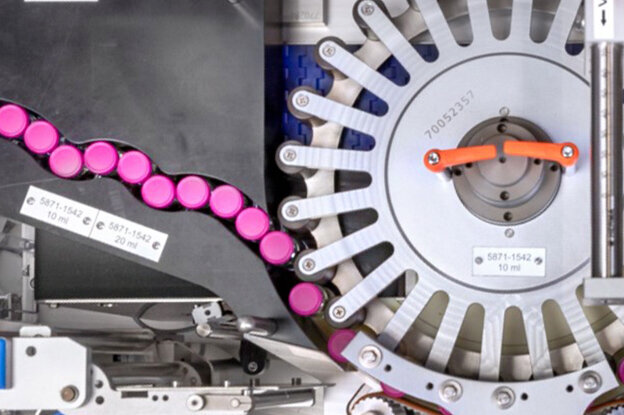 A person in protective gear is holding a syringe filled with a liquid, while in the background, a technical machine with colorful components is visible.