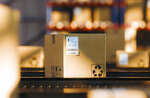 A brown shipping box with a label displaying recycling symbols and instructions is sitting on a conveyor belt in a warehouse.