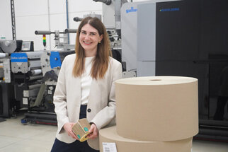 A woman stands next to large rolls of paper in a printing house, smiling and holding a small package in her hand.
