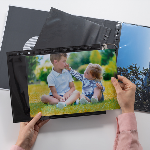 Eine Hand hält ein Foto von zwei Kindern, die im Freien auf dem Gras sitzen und miteinander interagieren, während ein Fotoalbum geöffnet ist.