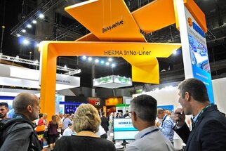 People are standing at a trade show booth named 'HERMA InNo-Liner,' which is adorned with a striking yellow banner.