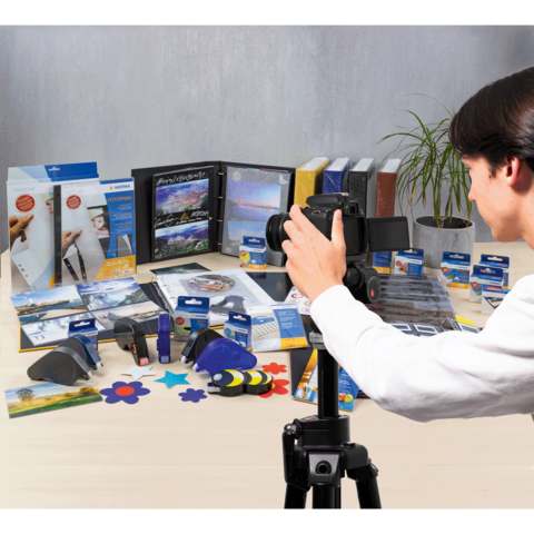 Eine Person fotografiert eine Vielzahl von Büro- und Bastelmaterialien, darunter Ordner, Kleber und Scheren, auf einem Tisch.