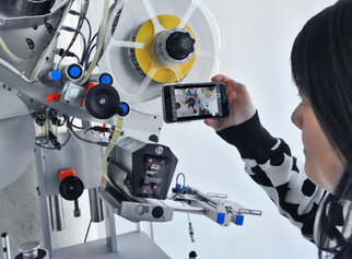 A person is holding a smartphone and taking a photo of a machine with various components and wheels.