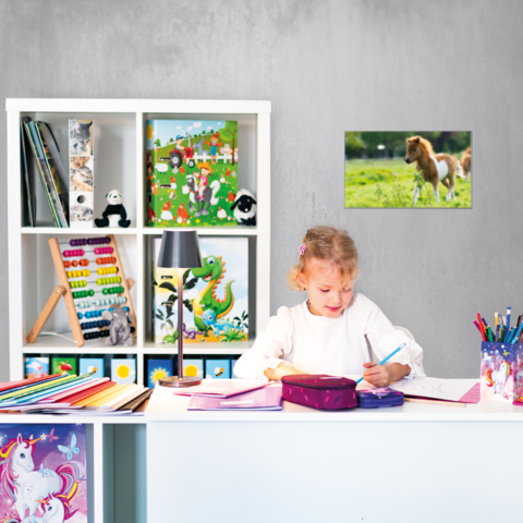 Ein Mädchen sitzt an einem Schreibtisch, schreibt in ein Heft und hat Schulmaterialien sowie bunte Papiere um sich herum.