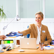 A woman in an office hands a man a document while sitting at a desk with a laptop and office supplies.