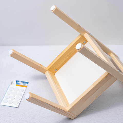 Umgedrehter Holzstuhl mit sichtbarem Sitz und einer Packung selbstklebender Polster auf einer grauen Oberfläche.