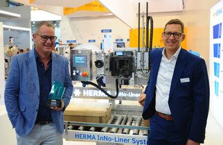 Two men are standing next to a HERMA InNo-Liner system, a machine for labeling, while one of them is holding a prize.