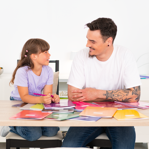 Ein Mann und ein Mädchen sitzen an einem Tisch, umgeben von Farbmustern und Materialien, während sie miteinander sprechen.
