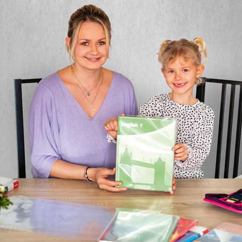 Eine Frau und ein Mädchen sitzen an einem Tisch und halten ein grünes Heft. Auf dem Tisch liegen verschiedene Schreibmaterialien und Blumen.