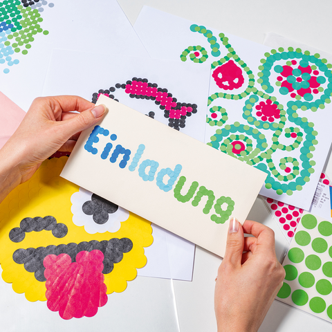 Hände halten eine Einladungskarte mit dem Wort "Einladung", umgeben von bunten Bastelmaterialien und Aufklebern auf einem Tisch.