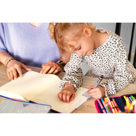 Ein kleines Mädchen schreibt in ein Notizbuch, während eine erwachsene Person daneben sitzt und zuschaut. Bunte Stifte liegen auf dem Tisch.
