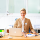 A woman with blonde hair is sitting at a desk, smiling, and wearing a beige blazer. In front of her is a laptop and some writing materials.