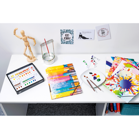 Ein Tisch mit Malutensilien: Aquarellfarben, Pinsel, ein Holzmodell und mehrere bunte Papierblätter. An der Wand hängen inspirierende Zitate.