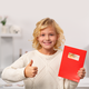 A girl with curly blonde hair is giving a thumbs up and holding a red envelope with a sticker in her hand.