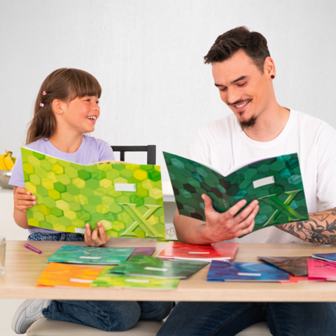 Ein Erwachsener und ein Kind sitzen an einem Tisch und betrachten bunte Hefte. Auf dem Tisch liegen mehrere Hefte in verschiedenen Farben.