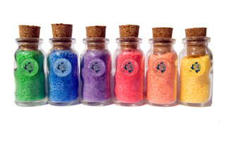 Six small glass bottles with corks, filled with colorful sand in the colors green, blue, purple, pink, orange, and yellow.