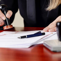 One hand is stamping documents on a table, while the other hand is holding a pen.