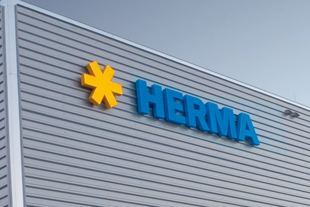 Facade of a modern building featuring the HERMA logo in blue lettering and a yellow symbol.