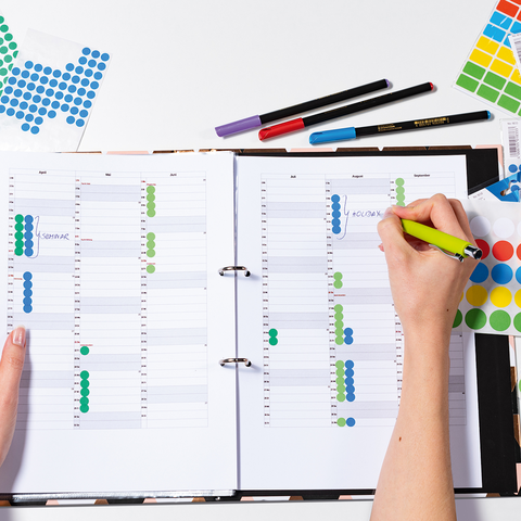 A opened planner with colorful stickers and handwritten notes. One hand is holding a green pen and marking appointments.