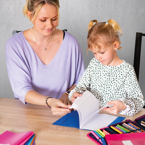 Eine Frau und ein kleines Mädchen sitzen an einem Tisch und blättern in einem Notizbuch. Bunte Stifte und ein Ordner liegen daneben.