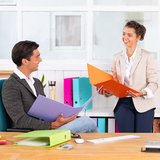Zwei Personen sitzen an einem Tisch und besprechen Dokumente. Eine Person hält orangefarbene Unterlagen, während die andere einen lila Ordner hält.