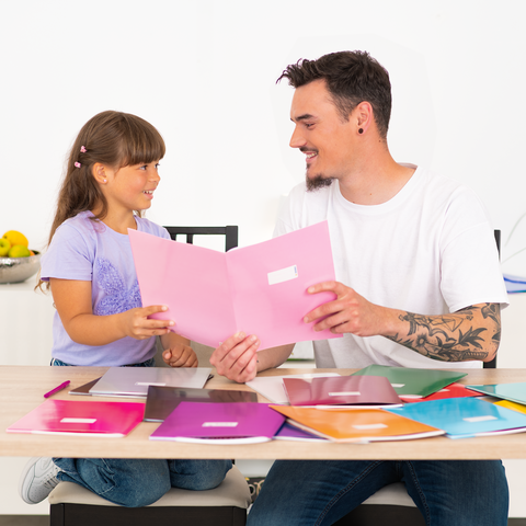 Ein Mann und ein Mädchen sitzen an einem Tisch und betrachten gemeinsam bunte Ordner. Der Tisch ist mit verschiedenen Ordnern bedeckt.